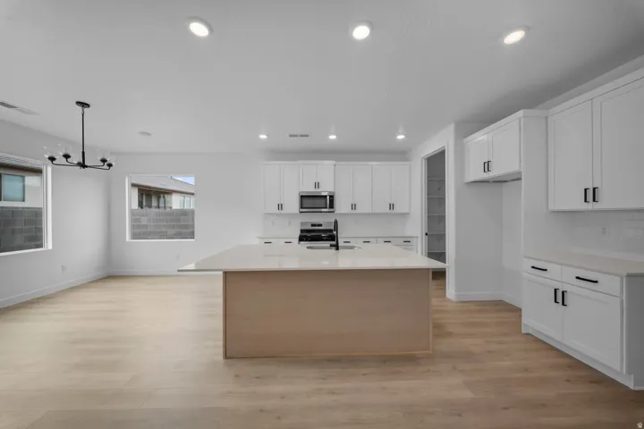Expansive open-concept living area featuring light wood-finish flooring, a contemporary chandelier, recessed lighting, and a kitchen with white cabinetry, stainless steel appliances, and a central island with a light-toned countertop