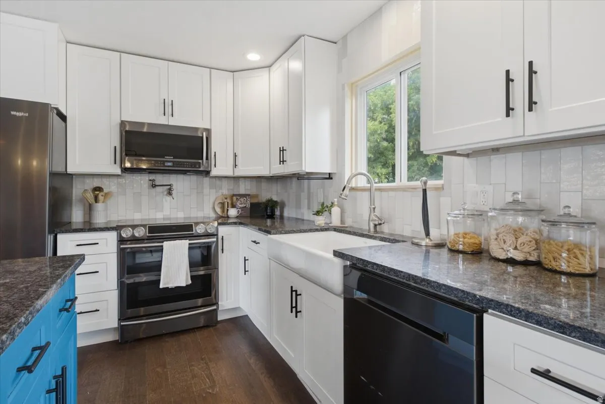 White shaker-style cabinetry with matte black hardware, stainless steel appliances, a farmhouse sink, dark countertops, and a vertical tile backsplash