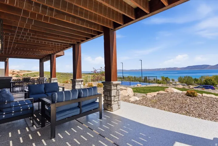 Covered patio with wood-beam ceiling and striped outdoor seating, overlooking manicured landscaping with native plantings, hardscaped pathways, and expansive water views of the lake and distant mountains beyond