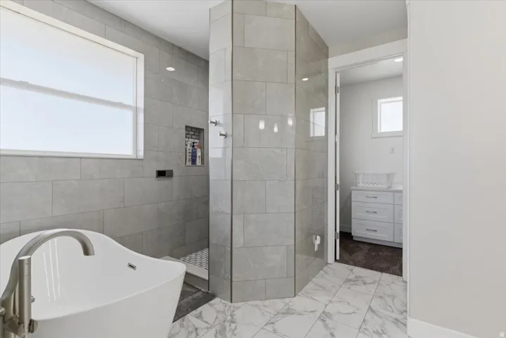 Freestanding soaking tub with floor-mounted faucet, walk-in shower with tiled walls and glass panel, recessed lighting, and white marble-patterned flooring