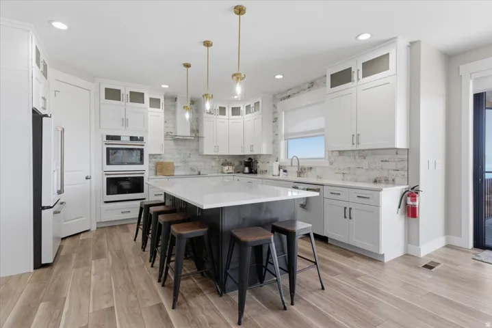 White shaker-style cabinetry with glass-front accents, stainless steel appliances, wood-finish flooring, a large kitchen island with seating, and a white tiled backsplash
