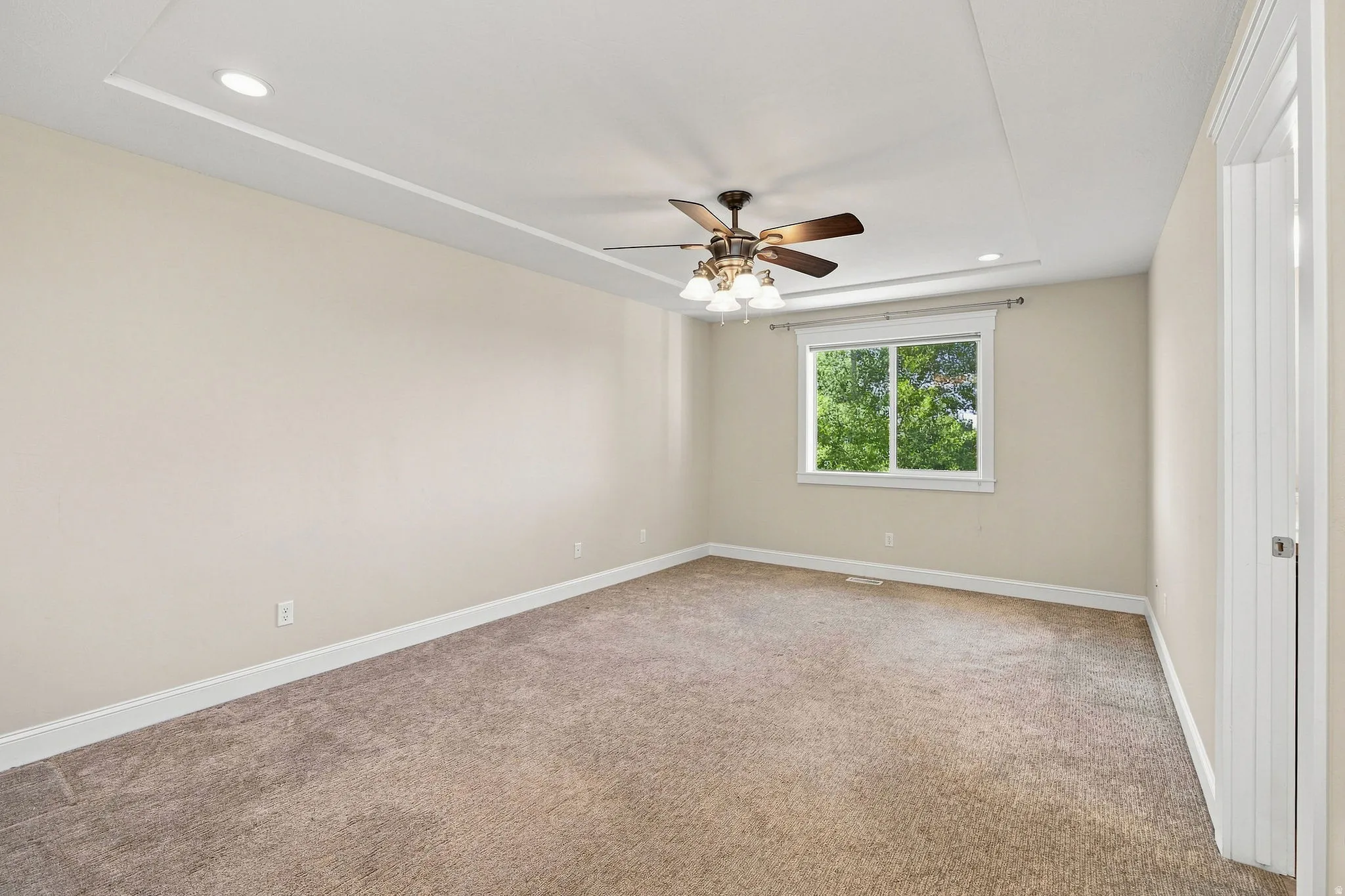 Primary bedroom. Tray ceiling with recessed lighting, ceiling fan with integrated light fixture, neutral carpet, white trim, and a window with exterior scenery