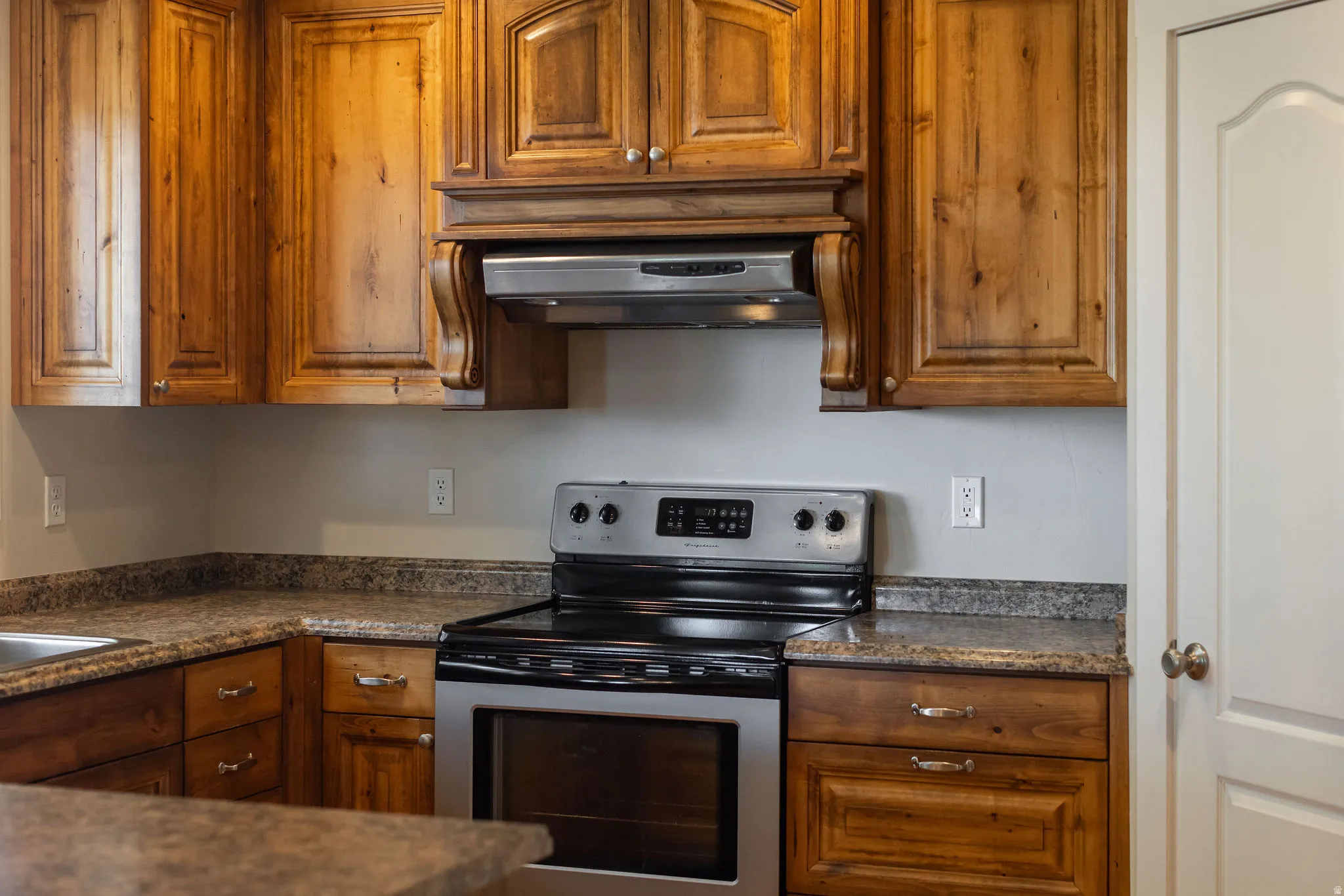 Wood-finish cabinetry with decorative hardware, stainless steel range with overhead hood, and light-toned countertop