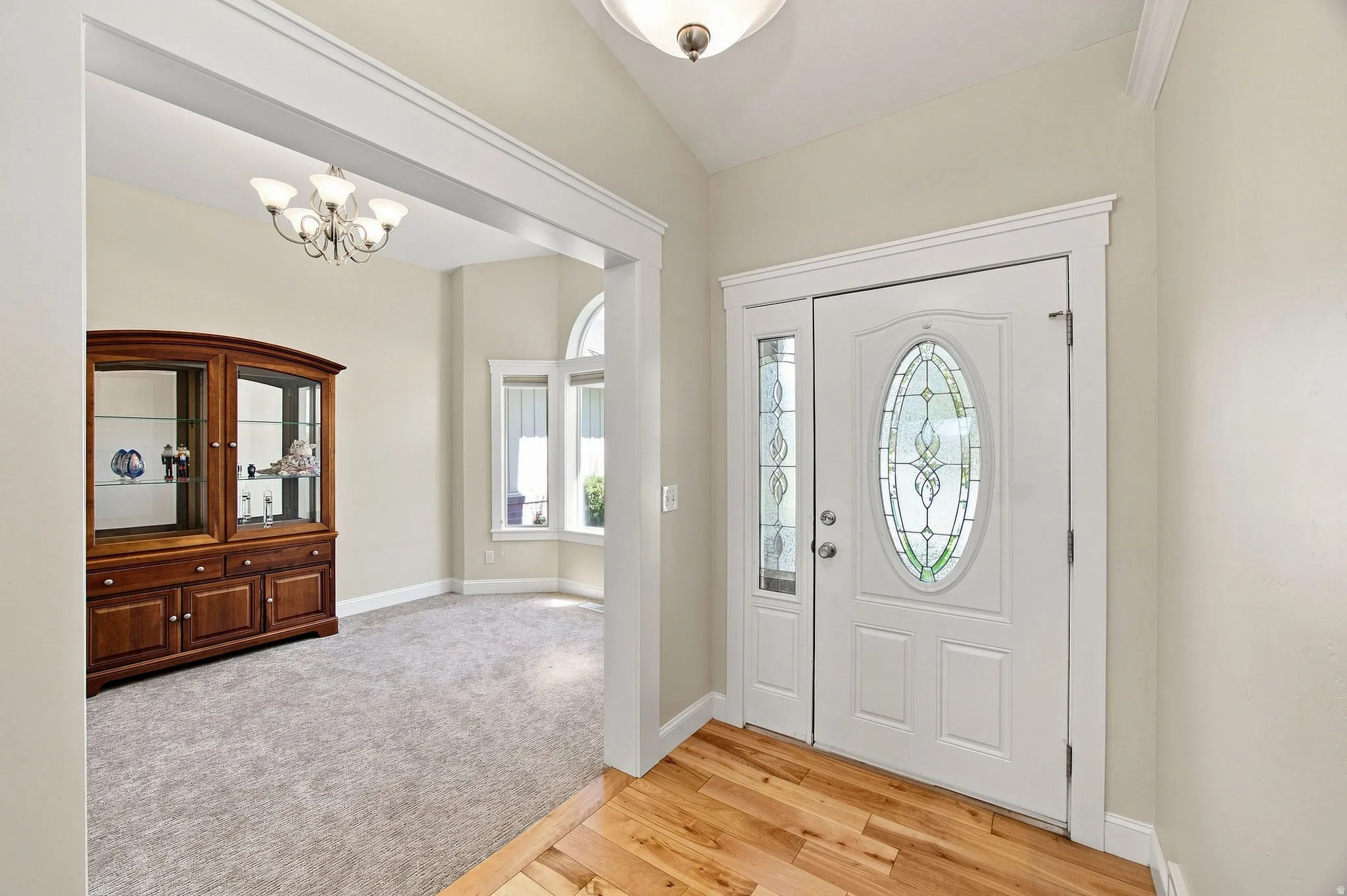Hardwood flooring entry with a white front door featuring decorative oval glass looking into front formal living room with carpet and bay windows.