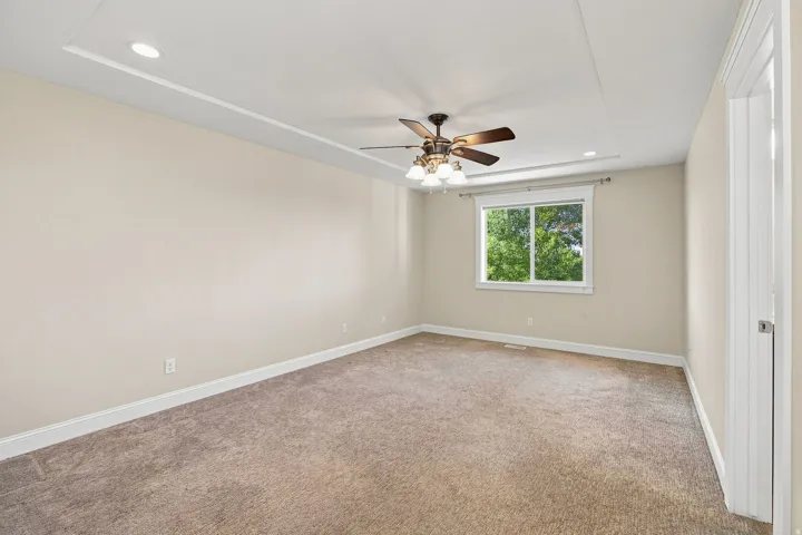 Primary bedroom. Tray ceiling with recessed lighting, ceiling fan with integrated light fixture, neutral carpet, white trim, and a window with exterior scenery