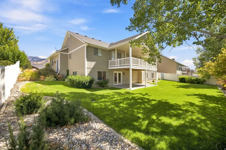 Expansive green lawn with mature landscaping, featuring a covered patio and an upper-level balcony with white railings