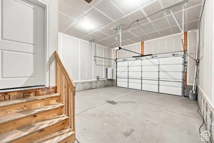 Concrete flooring, white panel garage door, automatic garage door opener, wood staircase with handrail, overhead lighting