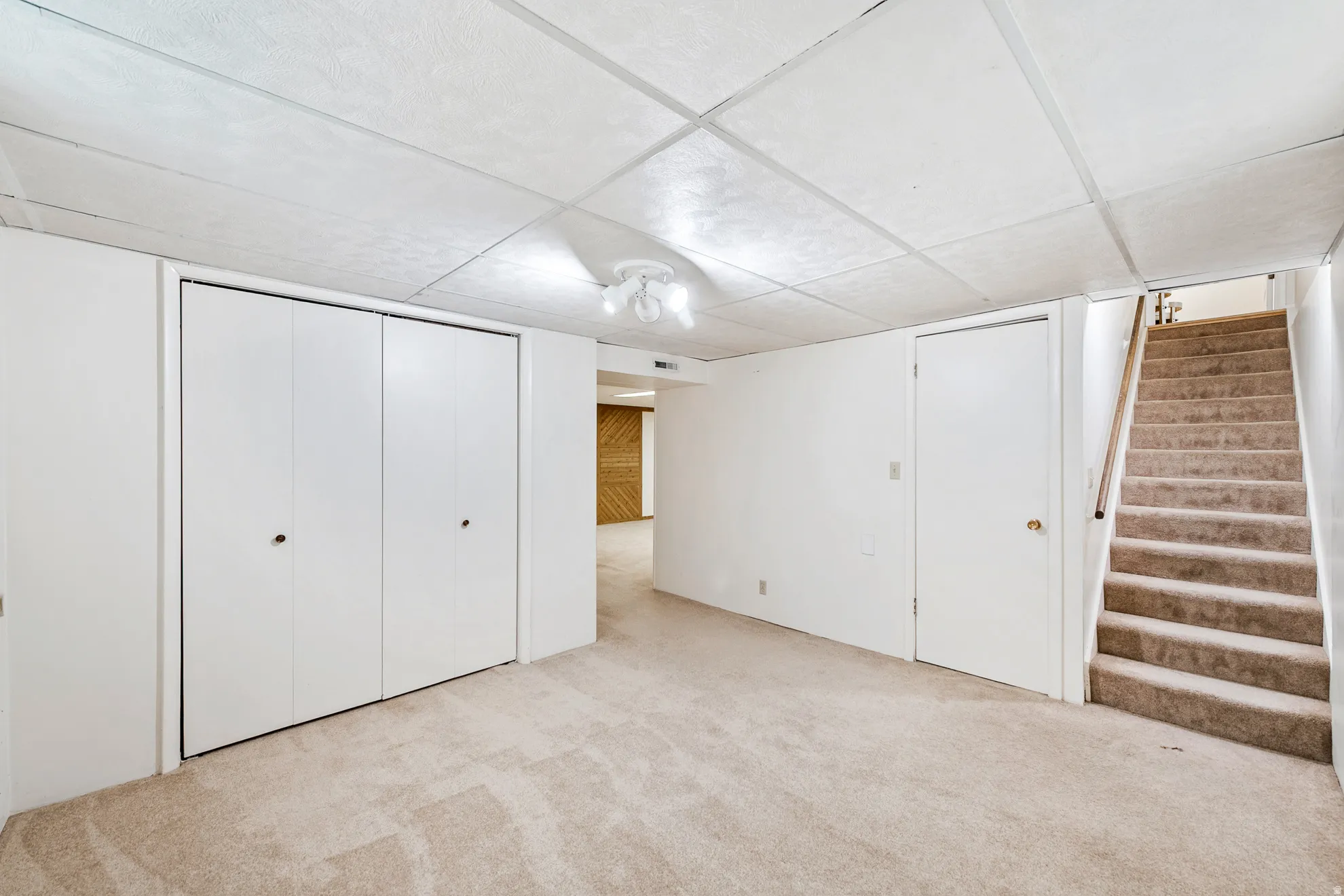 Carpeted flooring, drop ceiling, bi-fold closet doors, white interior door, and carpeted stairs with a wood handrail