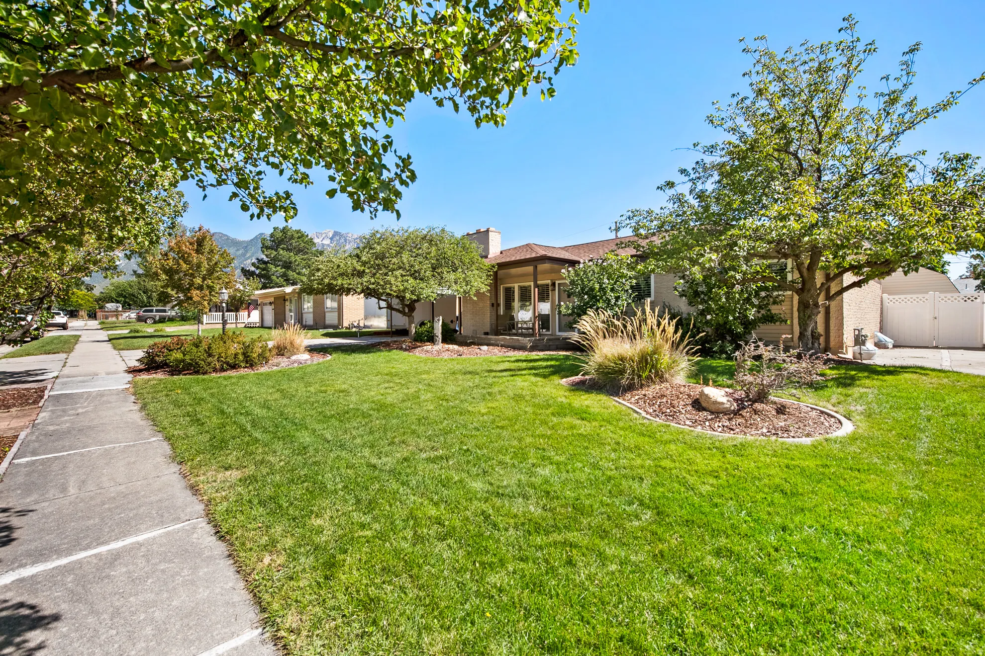 Spacious front yard featuring a manicured lawn, established landscaping, mature trees, and a concrete sidewalk