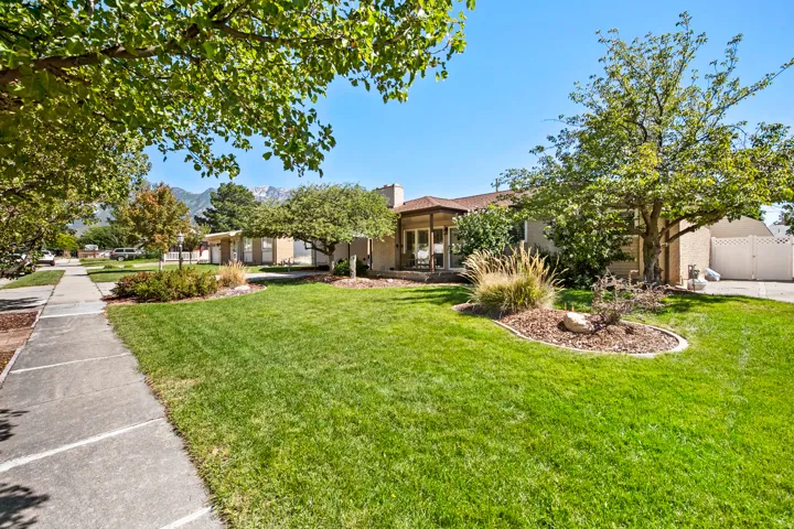 Spacious front yard featuring a manicured lawn, established landscaping, mature trees, and a concrete sidewalk