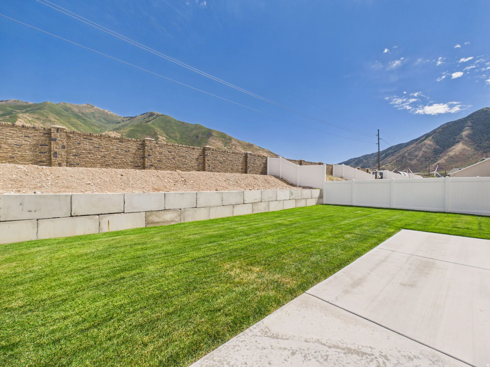 Expansive backyard featuring a lush lawn, concrete patio, a stone retaining wall, and a white privacy fence