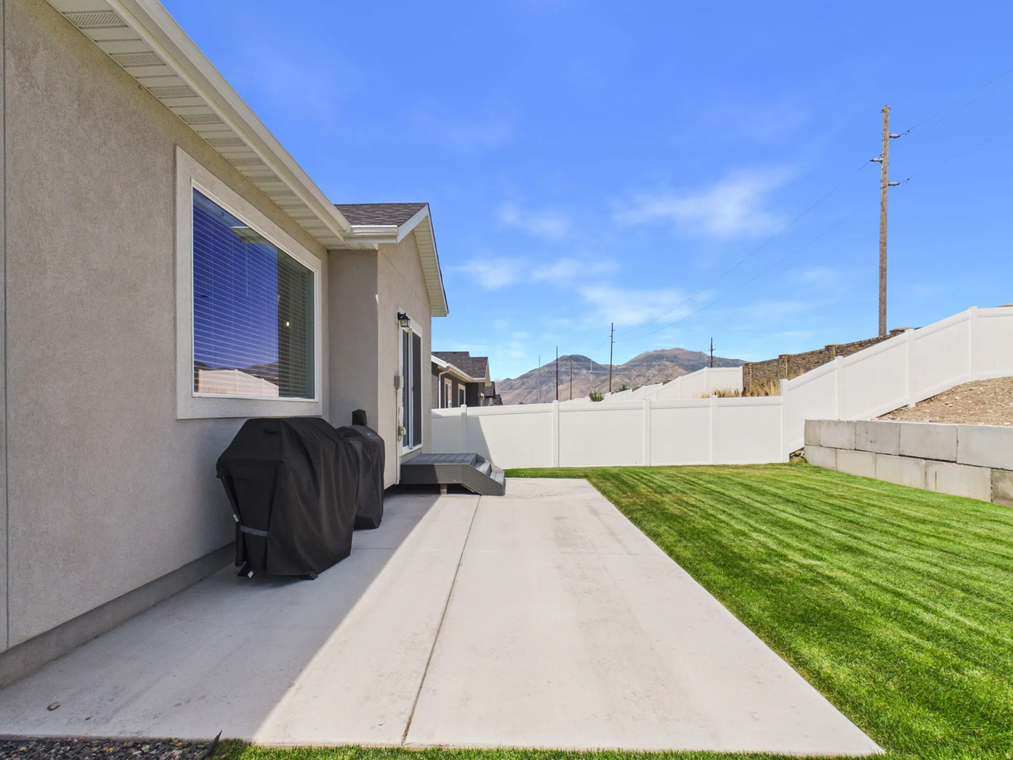 Expansive concrete patio with a manicured lawn, white vinyl fencing, and a concrete block retaining wall