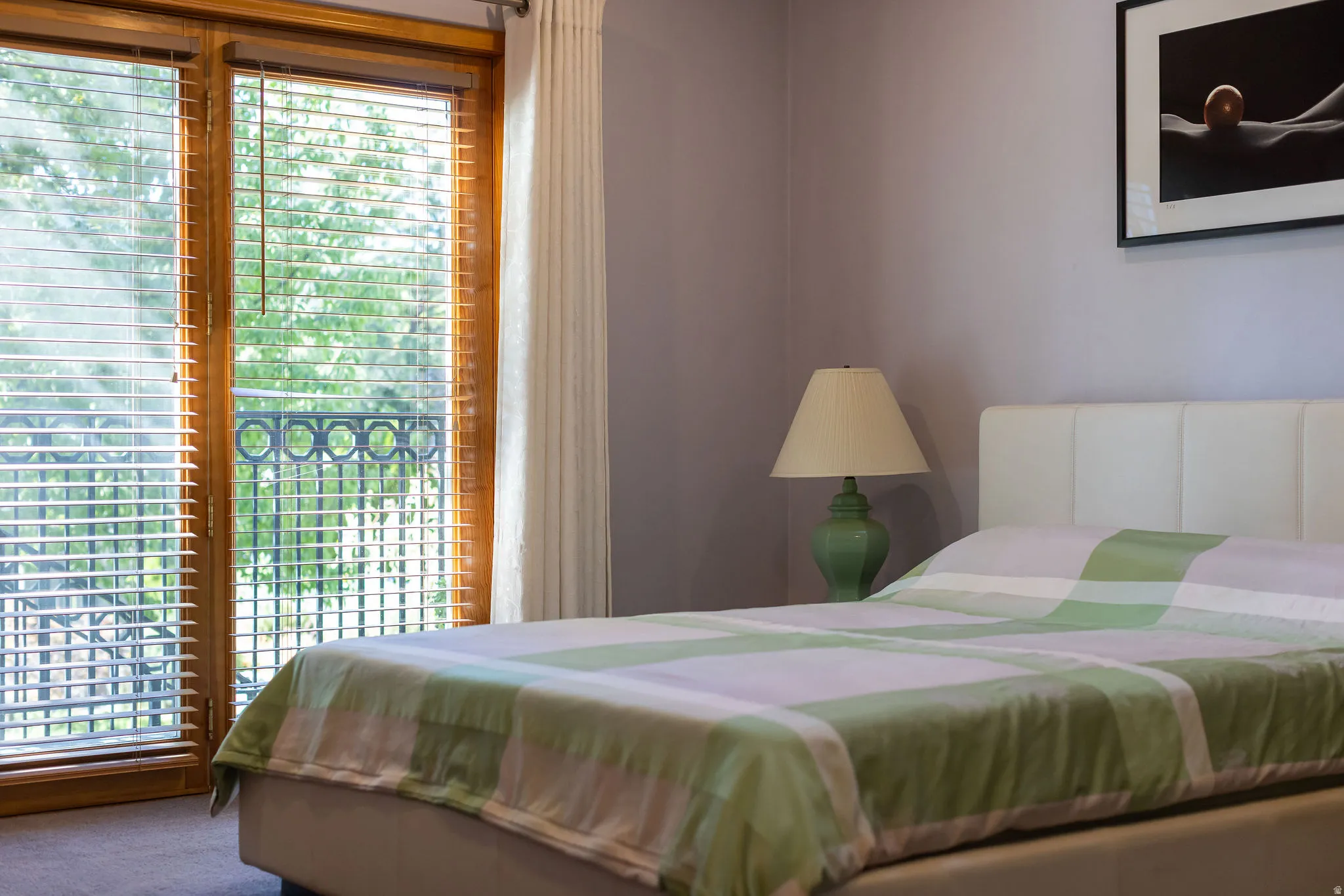 Wood-finish double doors with integrated blinds, iron balcony railing, upholstered headboard, and a bedside lamp with a fabric shade