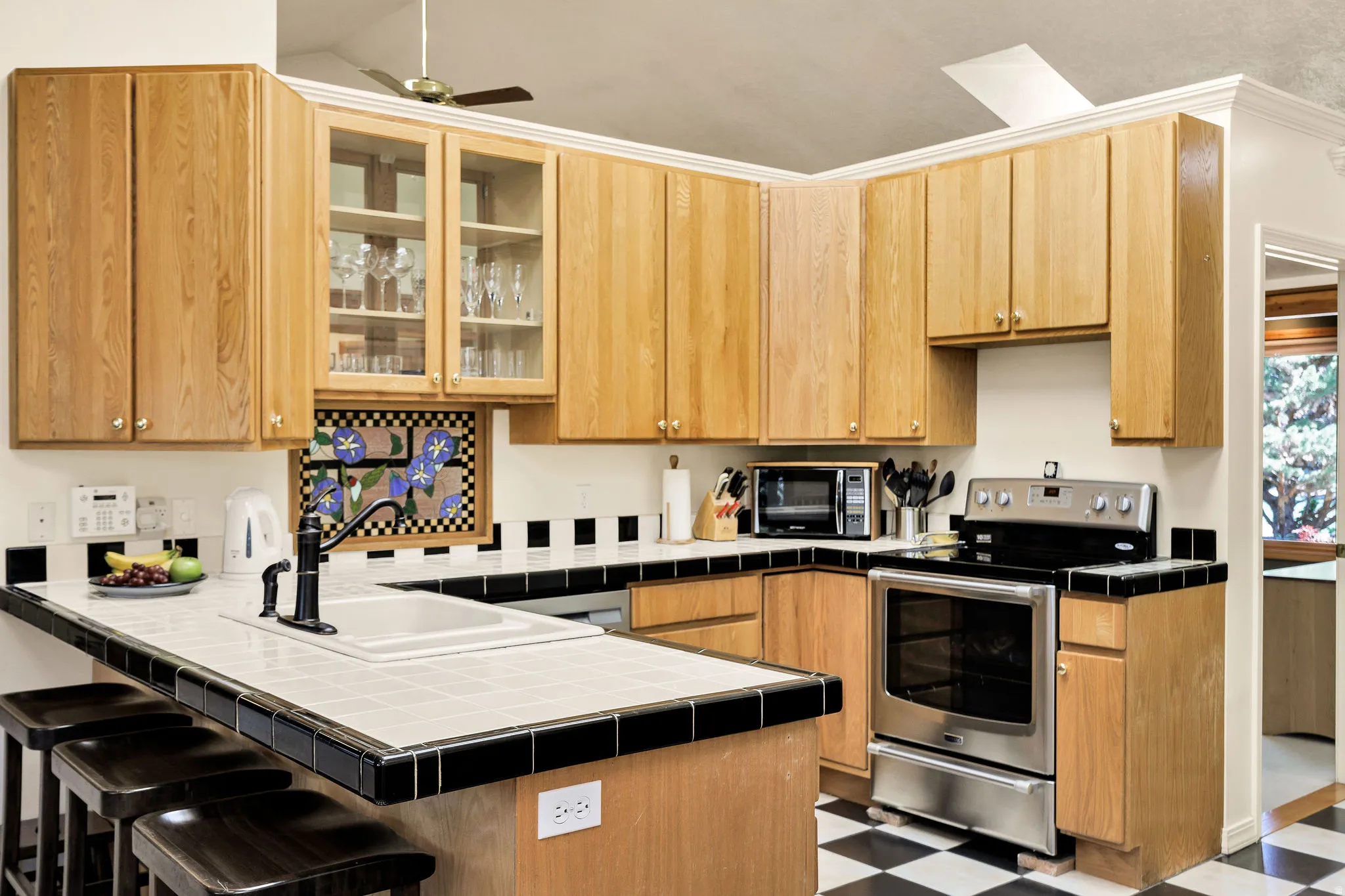 Kitchen featuring light wood cabinetry, a tiled breakfast bar, stainless steel range, and a black and white checkerboard floor