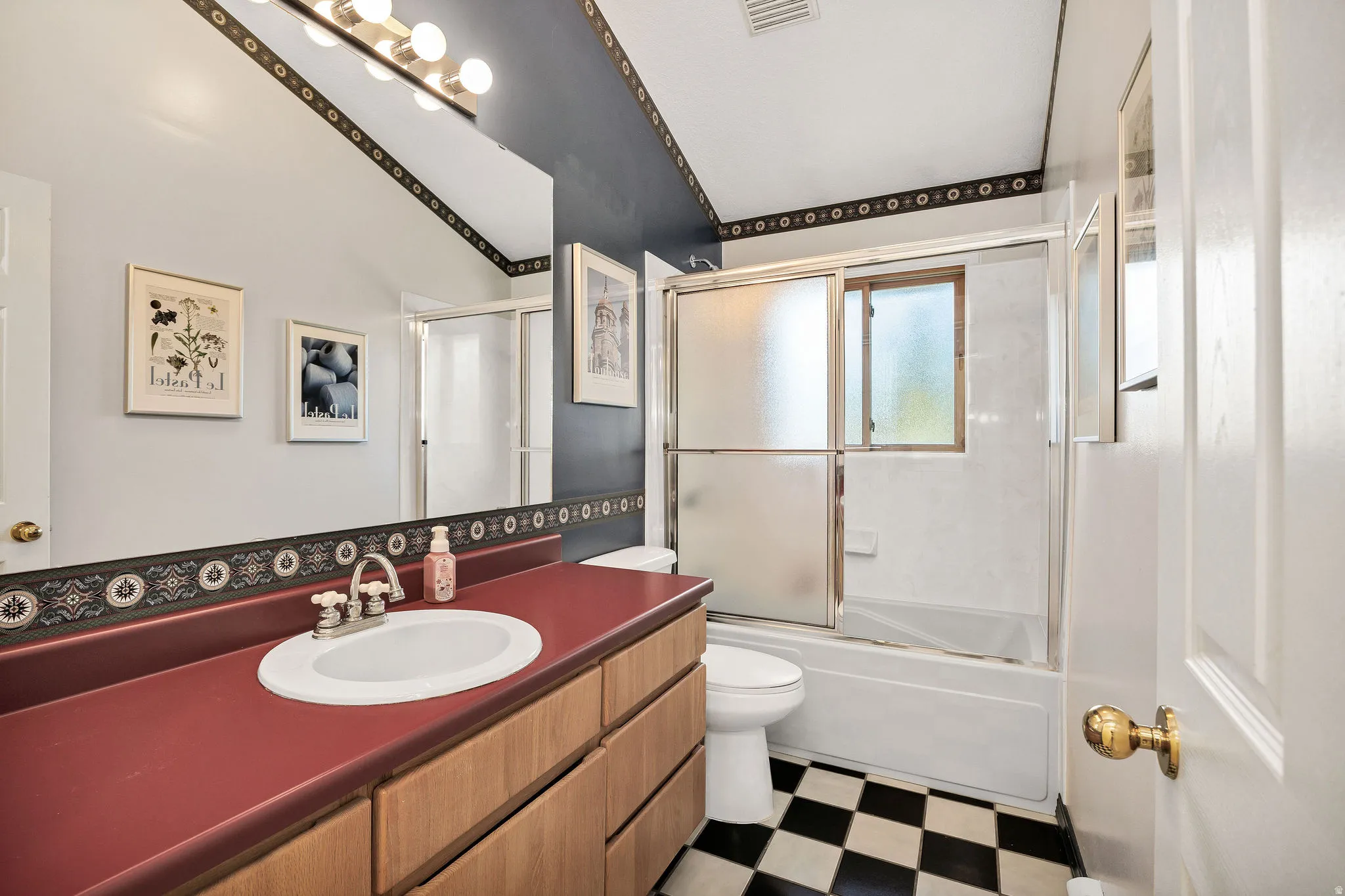 Bathroom featuring a wood-finish vanity with a red countertop and an integrated sink, a checkerboard tile floor, and a shower-tub combination with a sliding glass enclosure