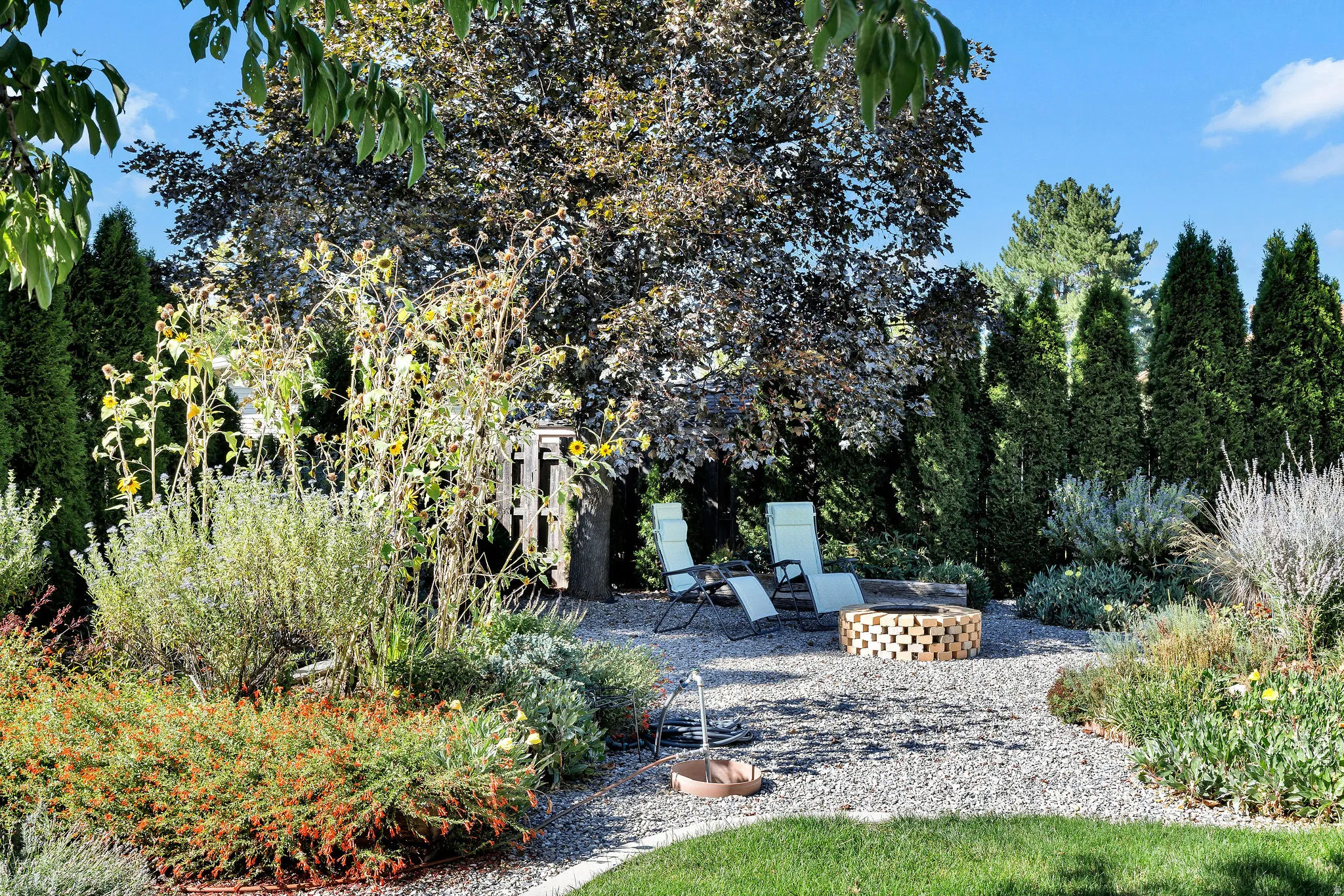 Landscaped backyard featuring a pea gravel patio, built-in fire pit, mature trees, and a variety of plantings