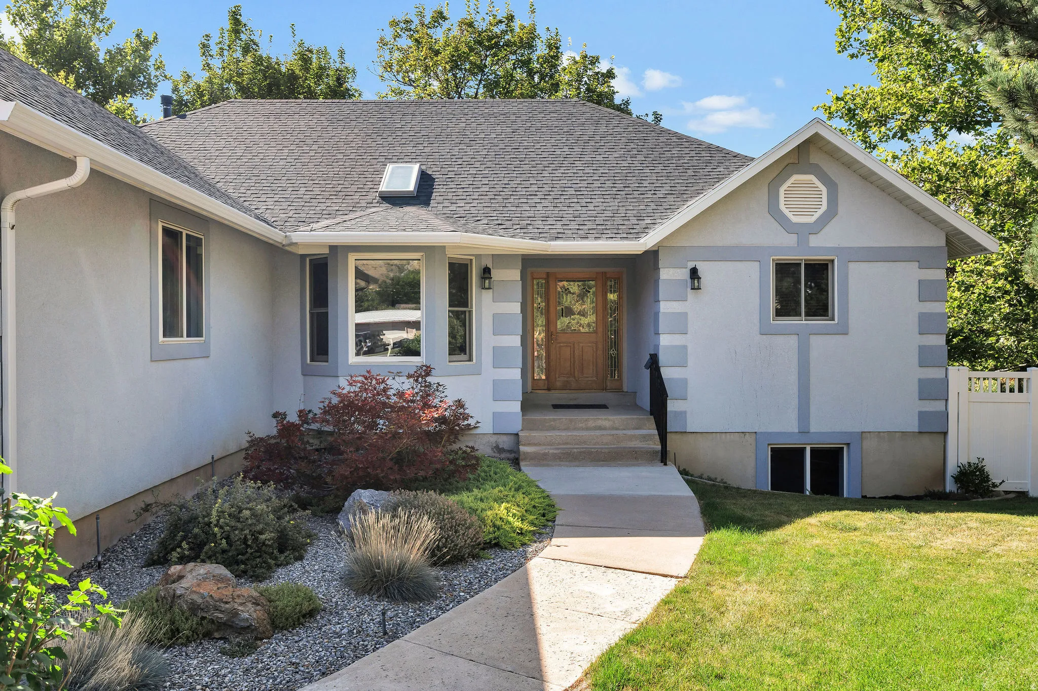 Wood-paneled entry door, mature landscaping, well-maintained asphalt shingle roof, integrated skylight, and concrete walkway