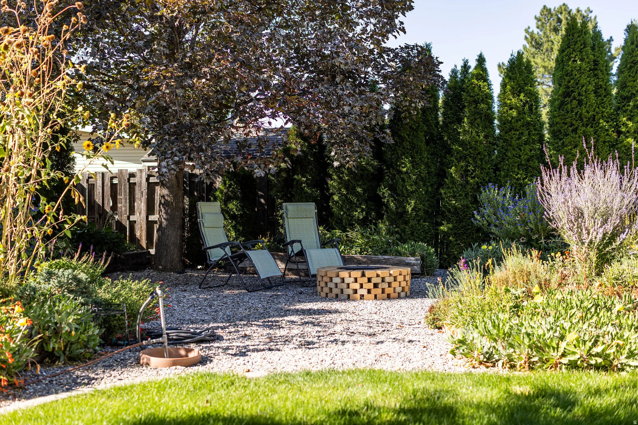 Gravel patio area featuring a built-in fire pit, mature shade trees, and privacy hedging