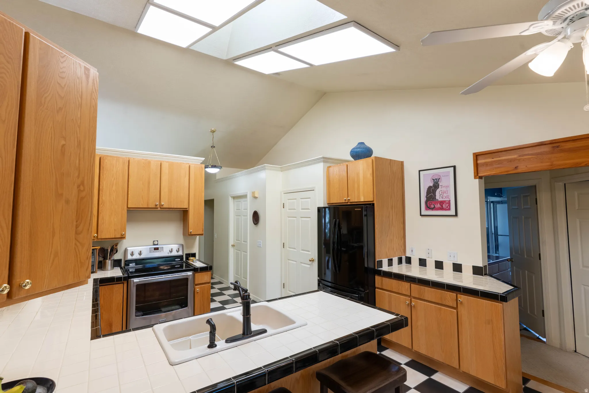 Vaulted ceiling with skylights, wood-finish cabinetry, tile countertops, black and white checkered flooring, and a ceiling fan