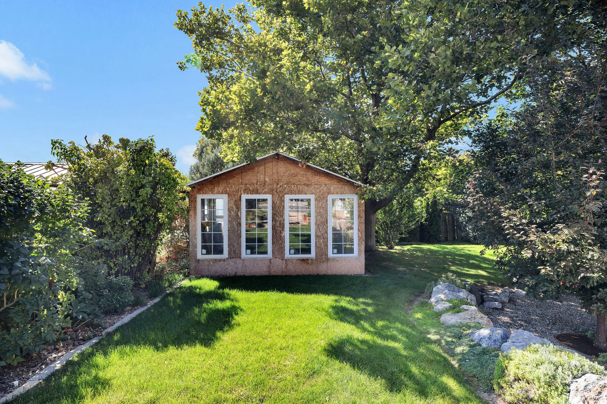 Expansive lawn featuring a detached accessory structure with multiple windows, mature trees, and natural rock landscaping