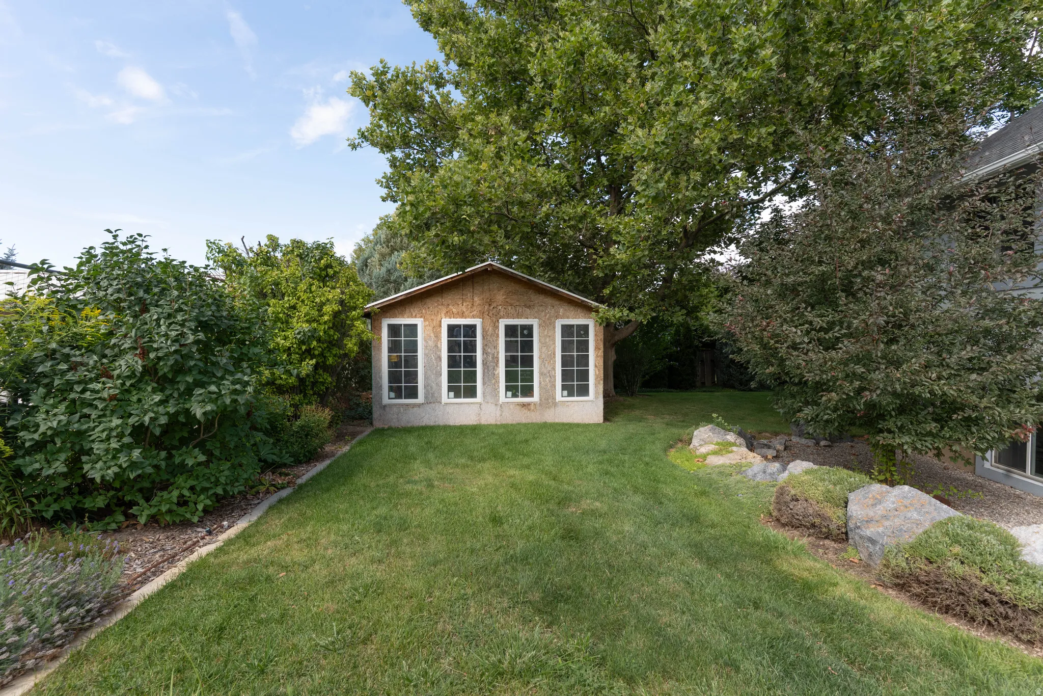 Landscaped backyard featuring a storage shed with multiple windows, mature trees, and natural rock accents