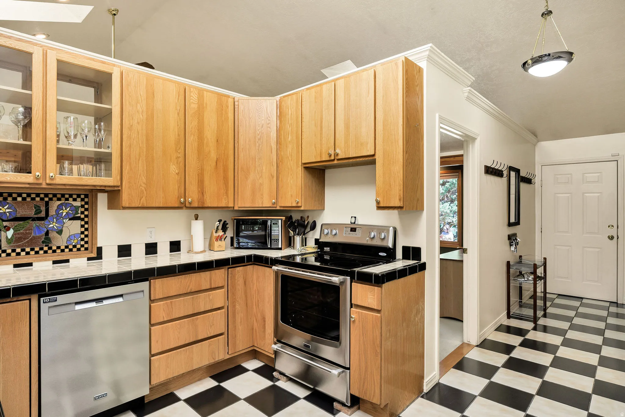 Kitchen featuring light wood cabinetry, checkered floor tiling, stainless steel appliances, and a vaulted ceiling