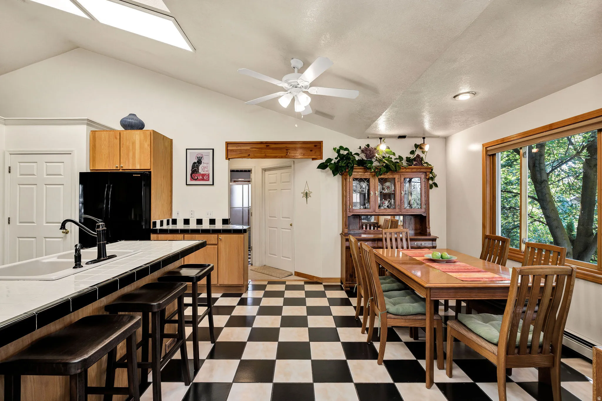 Vaulted ceiling with skylight, checkerboard tile flooring, wood-finish cabinetry, built-in kitchen island, and large window with exterior foliage