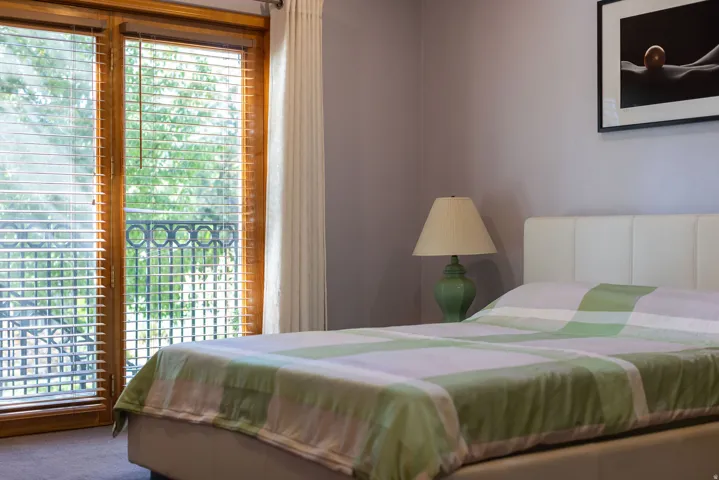 Wood-finish double doors with integrated blinds, iron balcony railing, upholstered headboard, and a bedside lamp with a fabric shade