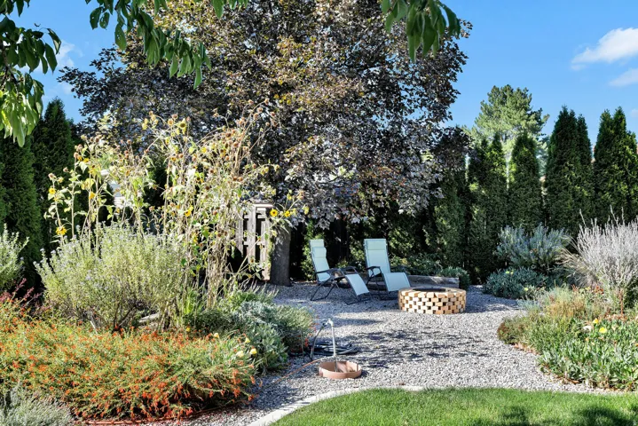 Landscaped backyard featuring a pea gravel patio, built-in fire pit, mature trees, and a variety of plantings
