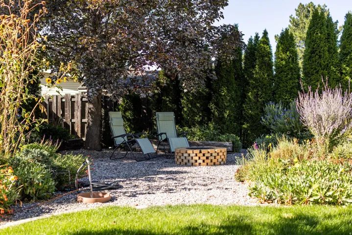 Gravel patio area featuring a built-in fire pit, mature shade trees, and privacy hedging