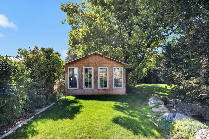 Expansive lawn featuring a detached accessory structure with multiple windows, mature trees, and natural rock landscaping