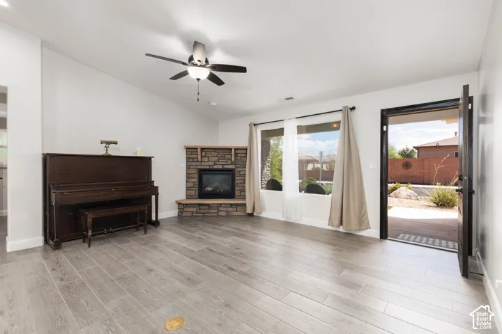 Vaulted ceiling with a ceiling fan, stone-clad fireplace, wood-finish flooring, and large windows