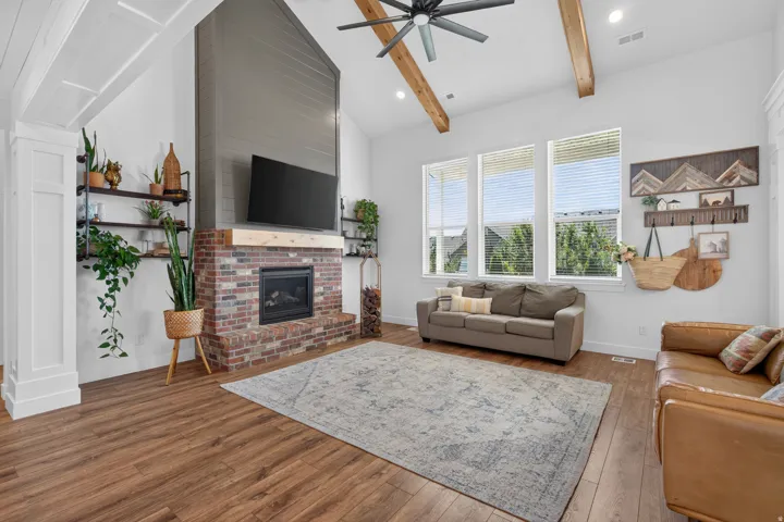 Vaulted ceiling with exposed wood beams, wood-finish flooring, brick-faced fireplace with wood mantel, large windows with blinds, built-in shelving