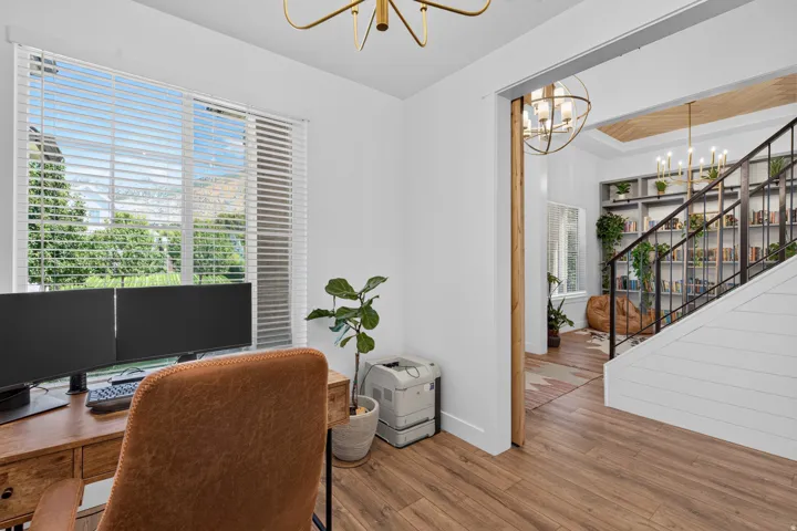 Wood-finish flooring throughout, bright window with horizontal blinds, overhead brass chandelier, open doorway with wood trim, and shiplap wall detail