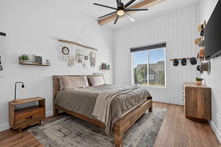 Vaulted ceiling with exposed wood-finish beams, ceiling fan, wide window with blinds, wood-finish flooring, and white vertical paneling
