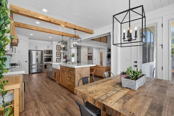 Wood-finish flooring throughout, white paneled ceilings with exposed wood beams, white kitchen cabinetry with stainless steel appliances, kitchen island with farmhouse sink, and a black metal chandelier
