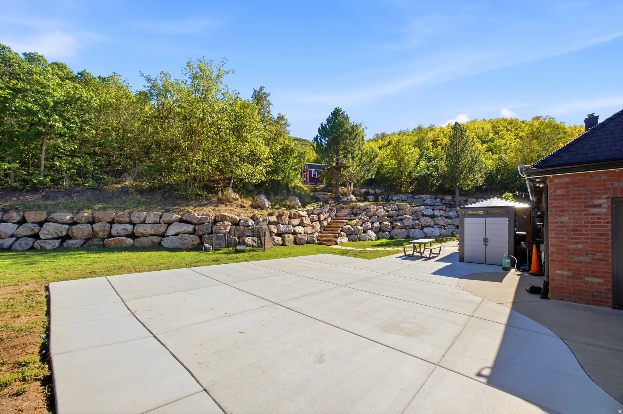 Expansive concrete patio with a tiered rock retaining wall and mature trees