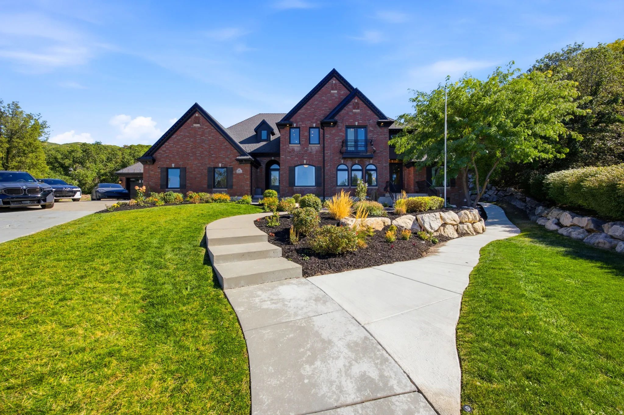 Brick exterior with black trim, concrete pathway, tiered landscaping with natural stone, mature trees, and a manicured lawn