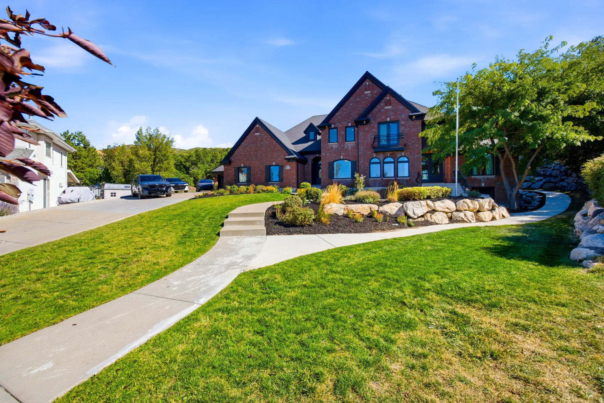 Brick exterior with dark trim accents, manicured lawn, stone retaining walls, mature landscaping, and a concrete pathway