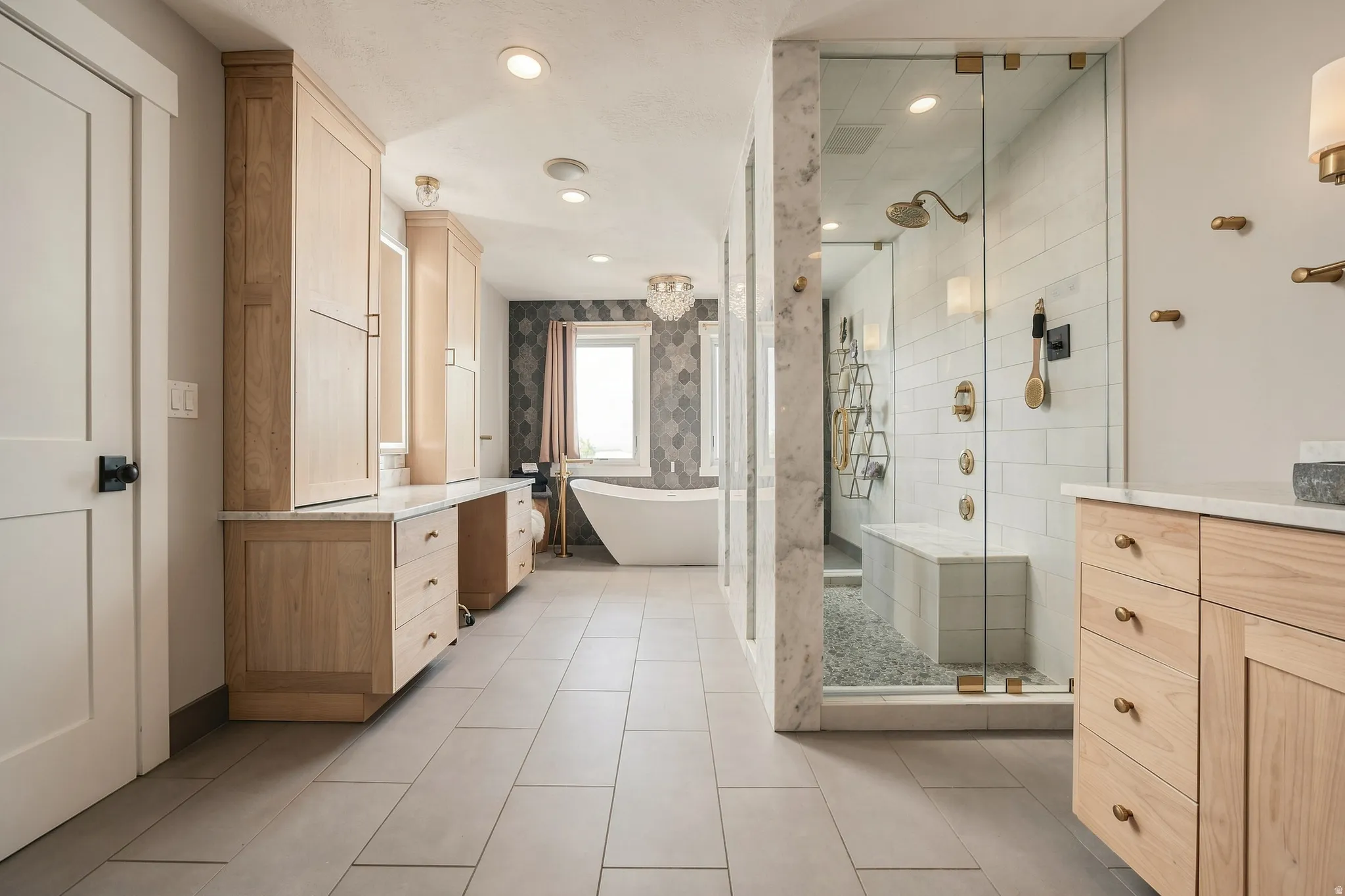 Dual light wood-finish vanities with stone countertops, a freestanding soaking tub, a spacious glass-enclosed shower with a built-in bench, and large format tile flooring