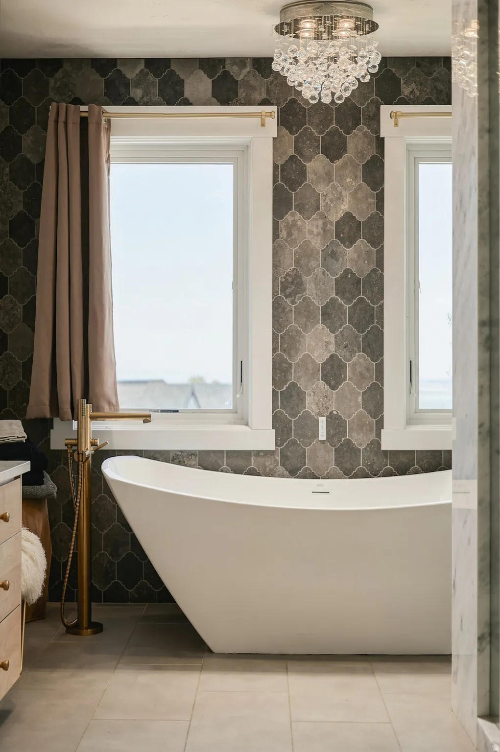 Freestanding soaking tub with a floor-mounted brass-finish faucet, hexagonal wall tiling, and a crystal-adorned flush mount light fixture