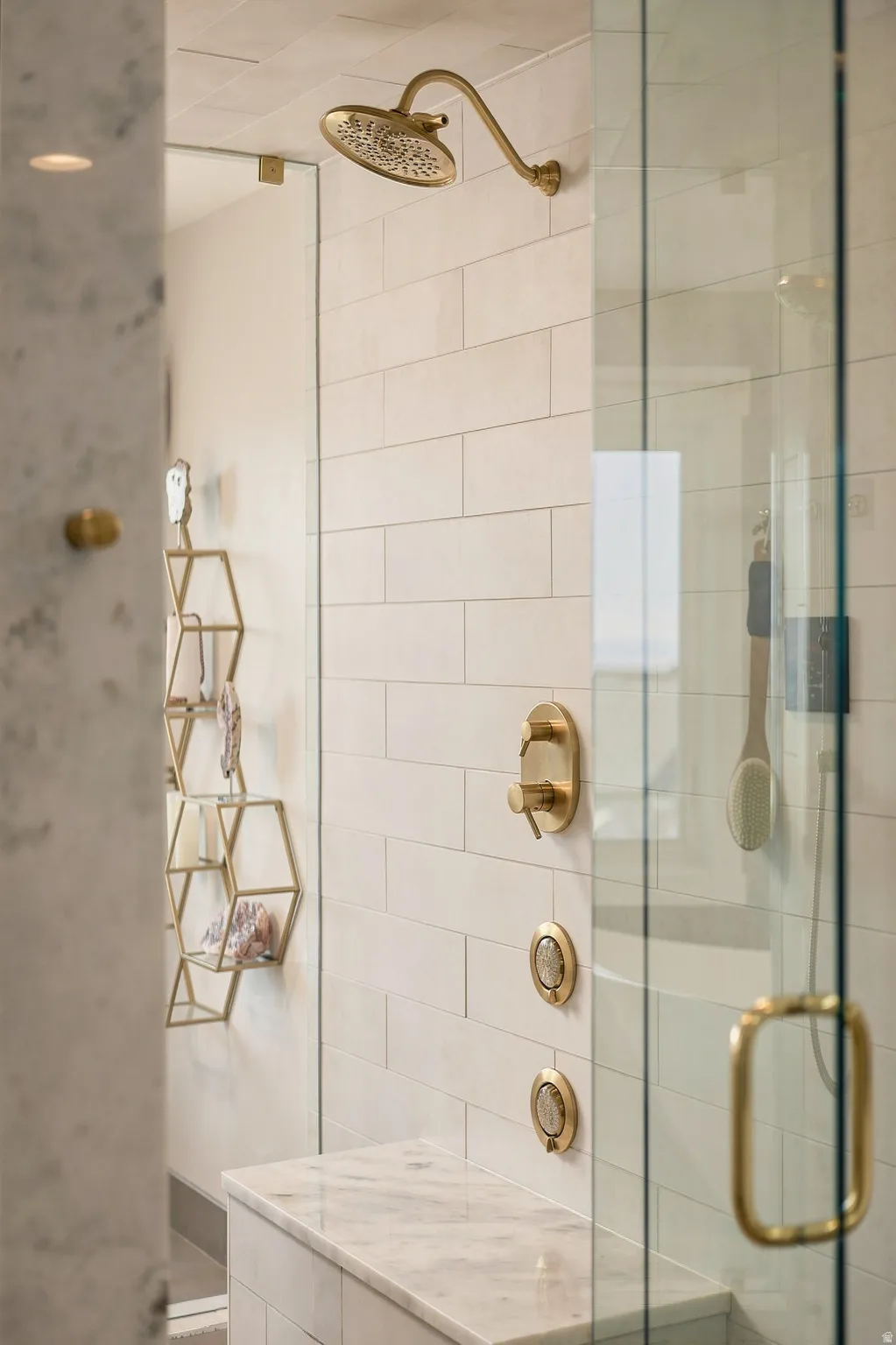 Shower with brass-finish fixtures, white subway tile walls, a built-in marble-finish bench, and a clear glass enclosure