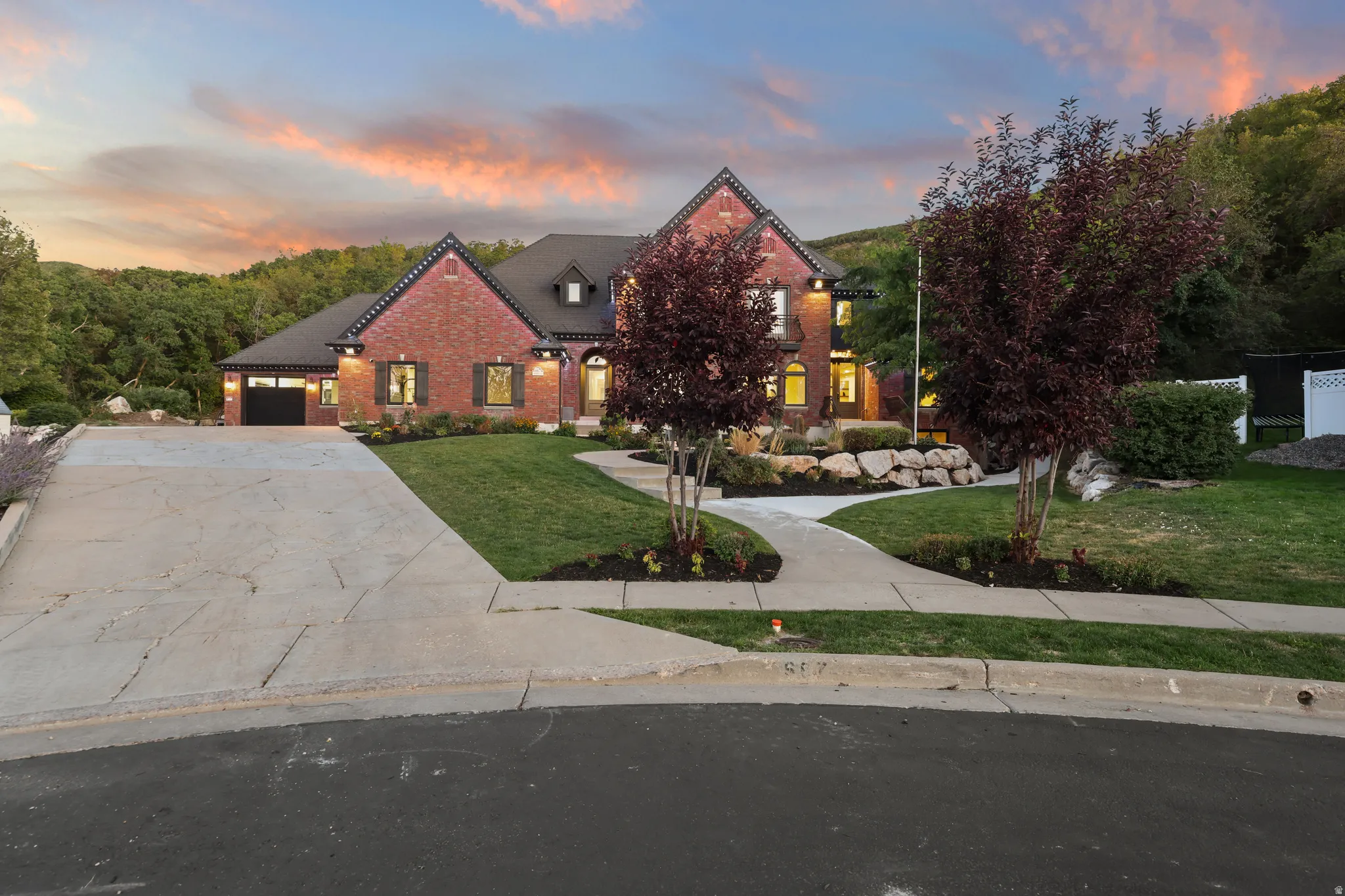 Brick exterior with black shutters, attached garage, concrete driveway, and mature landscaping