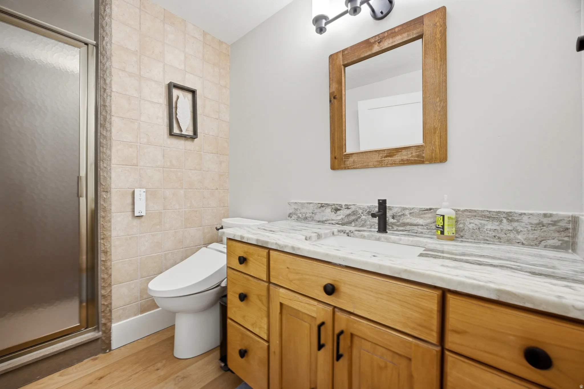Wood-finish flooring, tiled shower enclosure, natural wood vanity with black hardware, stone countertop, and rustic wood-framed mirror