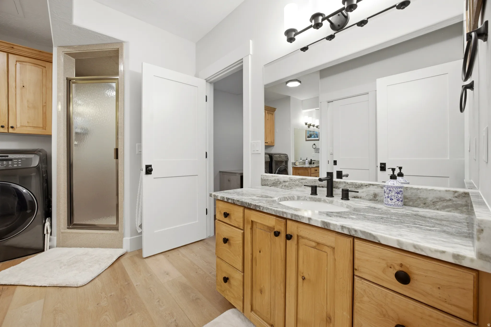 Light wood-finish flooring, wooden vanity with black hardware, stone-finish countertop, undermount sink with matte black fixtures, and a shower with a frosted glass door