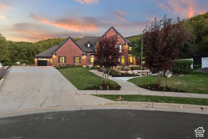 Brick exterior with black shutters, attached garage, concrete driveway, and mature landscaping