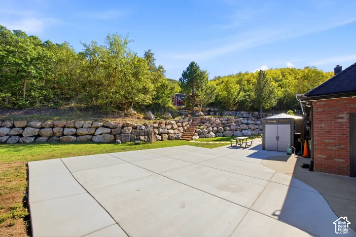 Expansive concrete patio with a tiered rock retaining wall and mature trees