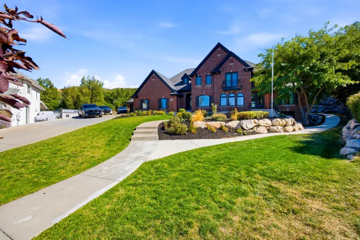 Brick exterior with dark trim accents, manicured lawn, stone retaining walls, mature landscaping, and a concrete pathway