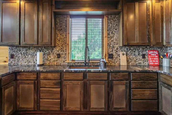 Dark wood-finish cabinetry, stone pebble backsplash, dark countertops, and an undermount sink with a dark-finish faucet