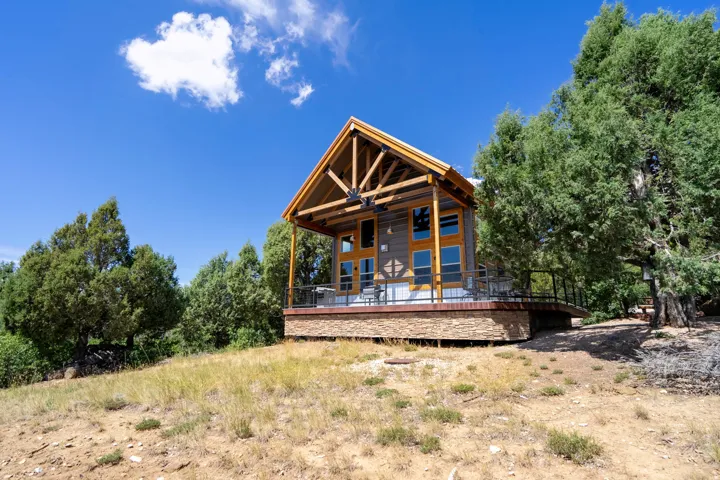 Gable roof design with exposed wood beams, composite wood siding, stone veneer foundation, expansive deck with metal railing, and surrounding mature trees